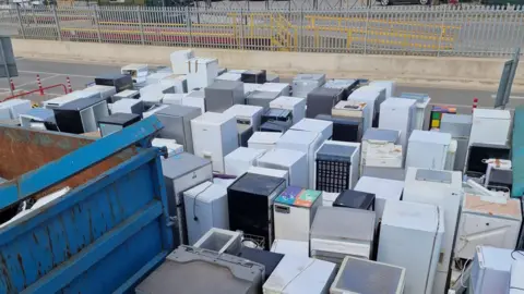 A large amount of fridges and freezers of varying colours and makes at a household recycling centre