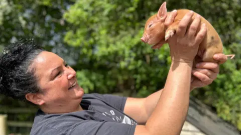 Kew Little Pigs Olivia Mikhail has dark hair and is holding a micro pig up in the air. She is smiling.