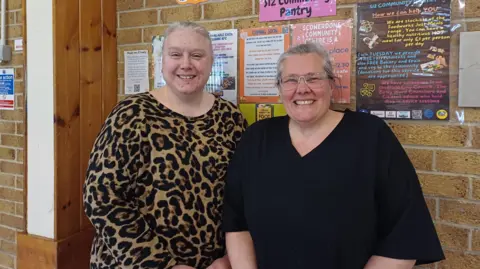 Julia Armstrong/LDRS The picture shows two women from the waist up.  Both are smiling.  One is wearing a leopard print top and has grey hair scraped back.  The other is wearing a black v-necked top and has clear framed glasses, she also has grey coloured hair scraped back from her face.  They are standing in front of a pale yellow brick wall which has a number of posters on it 