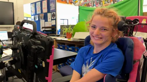 Phoebe sitting in a wheelchair looking towards the camera smiling. Her wheelchair is pink and she has a screen in front of her. She is in a classroom