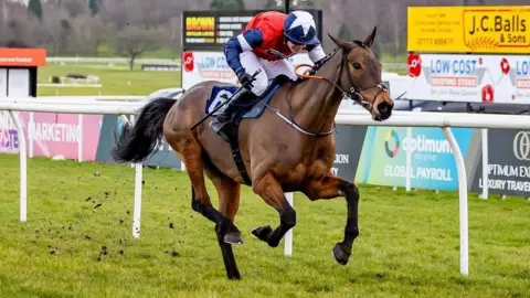 A jockey is riding a racehorse at speed on a grass racecourse. The horse is mid-stride with its front legs lifted high and its hind legs pushing off the ground, kicking up small clumps of turf behind it. The jockey is wearing a red, white and blue racing outfit and is leaning forward low over the saddle in a racing position. The number 6 is visible on the horse’s saddlecloth.