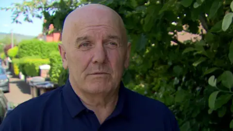 Kevin is standing outside. He is wearing a navy polo shirt. Leaves from a tree are next to him. Bushes are planted up the street. Cars are parked along the road. Brown wheelie bins are outside some houses. 