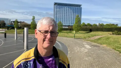 Amy Holmes/BBC A man in glasses wearing a purple sports jacket stands outdoors, with a modern glass building in the background.