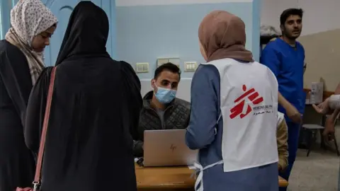 EPA Three Palestinian woman, one wearing an MSF tabard, gather round a table where a man wearing a surgical mask sits at a computer