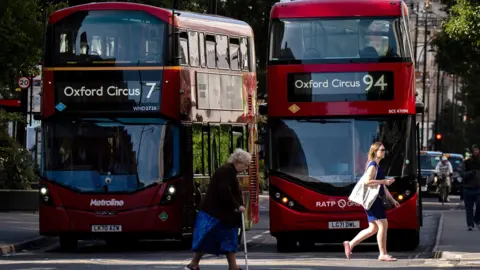 PA Media Two women, one with a walking stick, crossing the road while buses wait