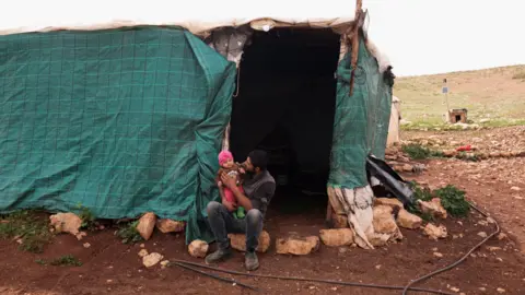 Reuters Suhaib Abu Kbash sits with his child on his lap outside their makeshift house in a Palestinian Bedouin community in the Jordan Valley, in the occupied West Bank (19 March 2026)