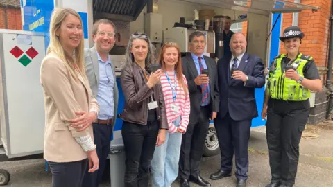 BBC Three men and four women standing in front of a mobile food van including a police officer and two men in suits, holding takeaway coffees.