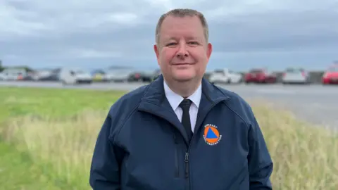 Dave Hodge is standing outside on grass with cars behind him smiling at the camera and wearing a waterproof jacket with a badge that reads 'Guernsey Civil Protection' on it.