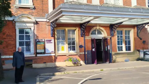 Harborough District Council Phil Knowles in a suit standing to the side of the front entrance to a red brick railway station with a purple door and an ornate porch