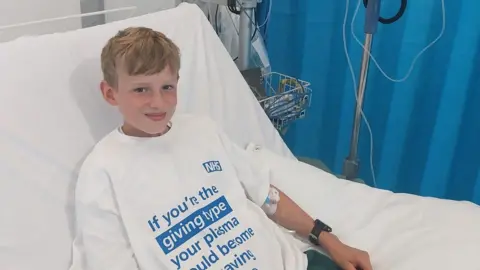George, who has blonde hair, smiles at the camera from a hospital bed. He is wearing a large white t-shirt with a message about giving plasma on it in blue lettering. 