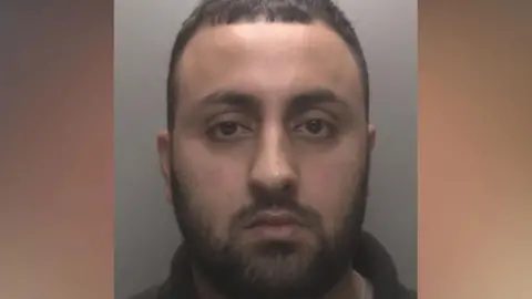 A man with short dark hair and a beard in custody photo.
