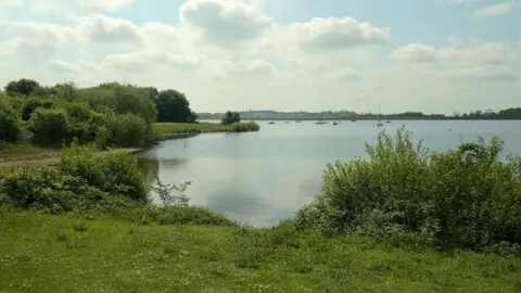 Carsington Water reservoir, which is owned by Severn Trent. The reservoir is a wide expanse of water with some boats visible in the far distance. The banks of the reservoir, with green grass and bushes, can be seen closer to the camera.