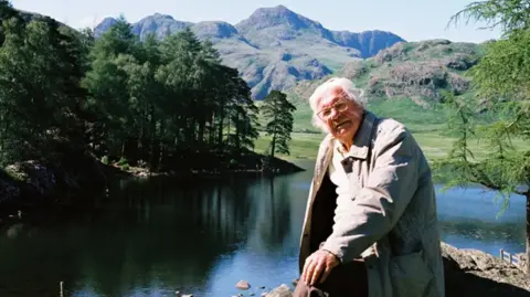 Chris Butterfield Alfred Wainwright, an older-looking man, wearing a beige outdoor jacket. He has shaggy white hair and wears glasses. He is standing in front of a lake with hills and trees in the background. 