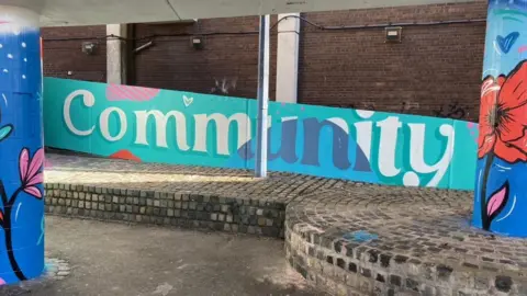 The word 'community' is painted in white and blue writing with turquoise and light blue behind it. To either side, two concrete pillars are painted blue with flowers and poppies.