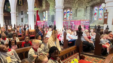 A large crowd of schoolchildren in grey uniforms, on wooden benches inside a cathedral, there are white beams and stone walls