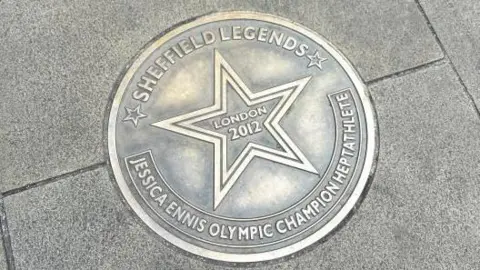 A gold-bronze coloured, circular metal plaque on the paving stone floor. Around the edge it reads "Sheffield Legends - Jessica Ennis Olympic champion heptathlete". In the centre is a star, with "London 2012" written in the middle of it.