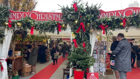 A gazebo with a red sign which says "Simply Christmas". There are people walking around in stalls and the carpet is red and grey. There are Christmas tree branches around. 