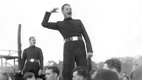 Getty Images A black and white photograph of a man giving a speech. He is standing on top of something and waving his fist. 