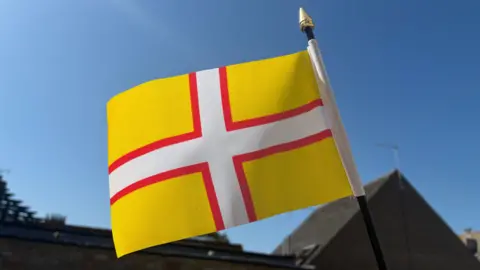 A huge yellow flag with a white cross, bordered in red, flies in front of a bright blue sky.