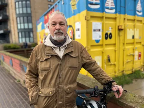 Image of Steven McCluskey, the founder of Bikes for Refugees. He is standing beside a bike in front of a blue and yellow container and is wearing a beige jacket with a cream hoodie underneath. His has short, grey hair and a grey moustache and goatee beard.