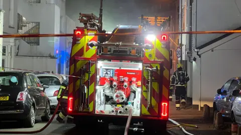 A picture of a fire engine parked on a residential street in St Helier on Tuesday evening. There are firefighters on the street and hoses all across the road. 