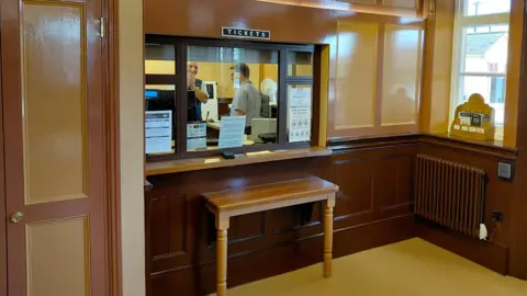 Daniel Mumby Wooden-paneled exterior of the ticket office. A black and gold sign reads "tickets" above the counter. The walls are painted in three shades of brown, in a classic style