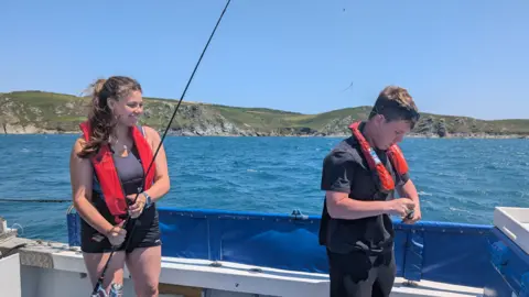 A young woman stands on the left holding a fishing rod. She is smiling because she just caught a fish. A teenage boy stands on the right removing the fish from the hook. They are both wearing red life jackets. 