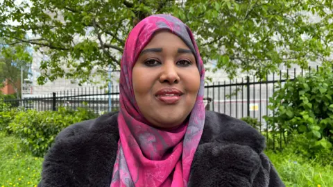 Samira is wearing a pink headscarf and black fur coat stood in a park with trees.