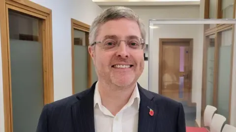 Philip Armstrong in a black jacket and white shirt smiles in a corridor with doors and chairs in the background