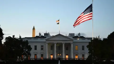 A view of the White House building from outside. An American flag flies in front of it, while the sun is setting.