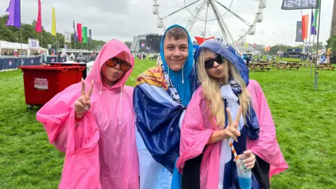 Three smiling fans at the TRNSMT festival, wearing pink and blue ponchos