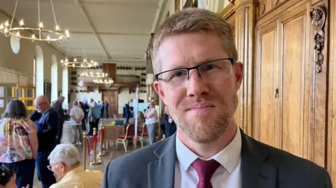 Mr Collins is wearing a grey suit, white shirt and maroon tie. He is stood inside a room with people in the background.