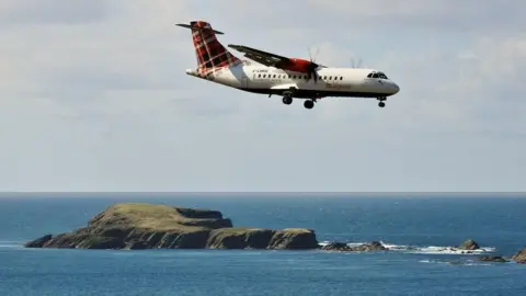 Loganair Mały samolot Loganair ze śmigłami w locie i kołami opuszczonymi do lądowania, z małą wyspą i błękitnym morzem w tle