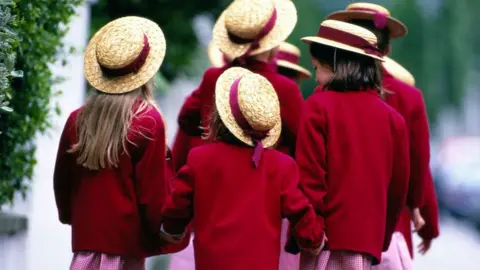 A group of young school girls wearing a private school uniform of red blazers and red and white check dresses and straw hats holding hands, facing away from the camera and toddling along the street to school