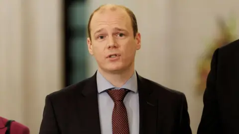 PA Media Gordon Lyons, wearing a blue shirt, dark red tie and black suit. 