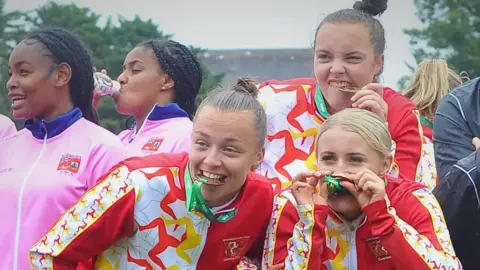 Rob Turville Three women from the Manx football team bite their bronze medals from the 2023 games in Guernsey. They wear bright kit, which is red, white and yellow with triskelions. They stand next to two other players from another team who wear pink.