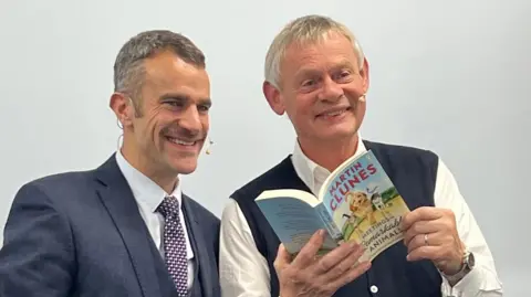 Pete Yendell Steve Harris and Martin Clunes smile for the cameras at the Dorchester Literary Festival. Steve Harris, on the left, is wearing a navy suit, white shirt and checkered tie. He has dark hair and a moustache. Martin Clunes is holding up a paperback copy of his book - Meeting Remarkable Animals.