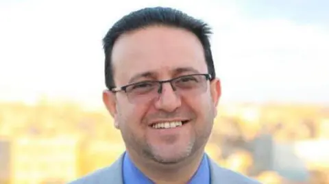 A man with short, dark hair and glasses looks into the camera, smiling, in an outdoor photograph