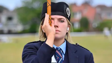 A young woman is holding a baton in a vertical position with one hand, which is at eye level. She has a cap on with a medal attached. He's wearing a navy jacket with lapels on her shoulders and a blue shirt and checked tie. 