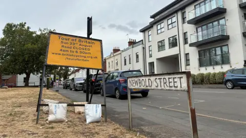 A close-up of a small white street sign which says Newbold Street. To its left is a large yellow road sign which says Tour of Britain, with information about the road being closed. Cars can be seen parked along the street and a large white building is on the other side of the road.