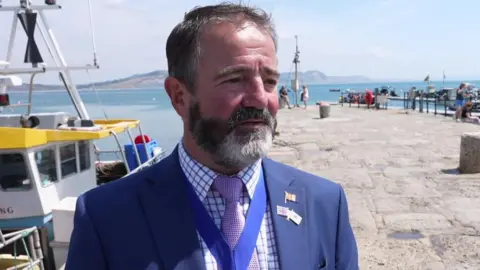 Phil Street is wearing a blue suit with a lilac tie. He has short brown and grey hair and a brown and grey beard. He is standing in Lyme Regis harbour.