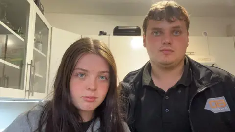Harvey Brett and Maya Port from Saltash stood in their kitchen and are looking straight at the camera with stern faces. There are a number of white kitchen units behind them, two of which have glass fronts. 