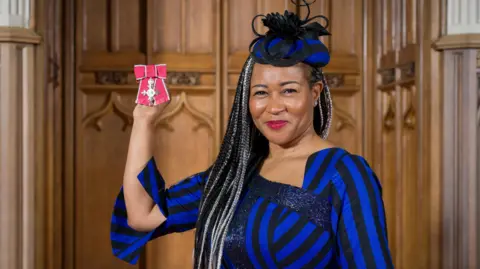 British Ceremonial Arts Limited Shalom Lloyd, holding up an MBE, wearing a blue and black hat and matching outfit, she is smiling and looking at the camera