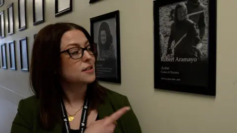 BBC A woman stands in a school corridor lined with framed photographs displayed in neat rows along the wall. She has dark-brown, shoulder-length hair and is wearing glasses, a gold necklace and a green jacket with a lanyard. She is pointing towards one of the framed portraits beside them. It shows an actor with long hair holding a sword in the TV show Game of Thrones.