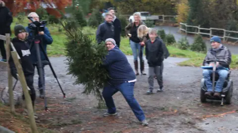 A person wearing blue trousers and blue jeans with a grey hat. They are leaning back, getting ready to throw a green Christmas tree. They are outside and surrounded by eight people watching.