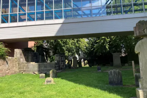 Gravestones in a grassy area at Greyfriars. There are trees at the back of the graveyard and the area is dominated by a glass and metal walkway.