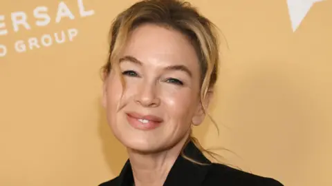 A woman with blonde hair poses on the red carpet in front of a yellow advertising sign. She is wearing a black jacket and is visible from the neck up. 