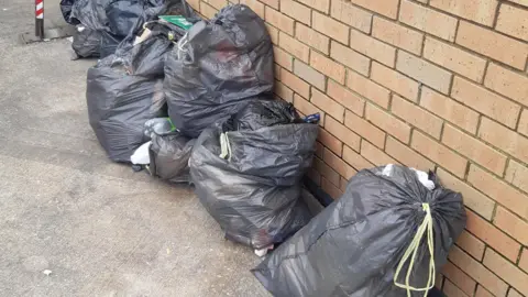 A large number of bags of rubbish, outside, by a brick wall. The bags are lying on the ground and others are all stacked up. 