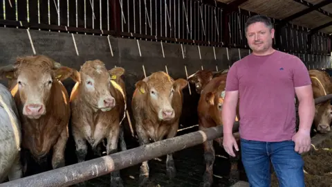 Jonny Tuft stands in a cattle shed, he is wearing a dark pink T-shirt and blue jeans. He has short black hair. To his right, behind a metal bar, are a row of cows looking towards the camera.