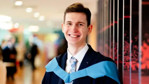 Open university Rainers is wearing a dark blaze, white shirts ad colourful tie, and across his chest and draped from his shoulders is a blue piece of material traditionally worn by people on graduation day.